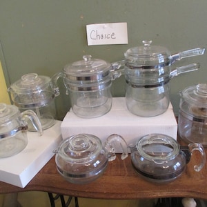 May include: A collection of seven vintage glass coffee percolators with silver accents. The percolators are arranged on a white surface and a wooden table.