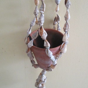 May include: A hanging terracotta planter adorned with strands of seashells. The planter is suspended by multiple shell-adorned cords. The shells are primarily beige and white, adding a natural, coastal aesthetic. The planter is empty.