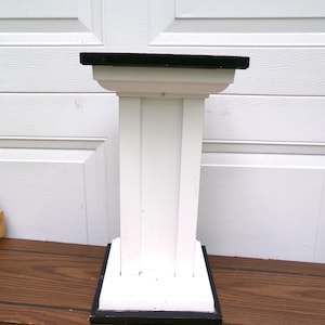 May include: A white pedestal stand featuring a square black top and a matching square black base. The central column is constructed with vertical white panels, and the structure includes a flared support section directly beneath the black top surface.