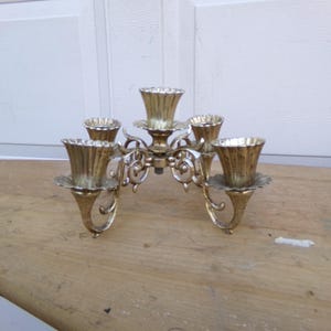 May include: A vintage brass candelabra with five candle holders. The candelabra has a decorative, ornate design with curved arms and a central column. The candle holders have a fluted design. The candelabra is on a wooden surface.