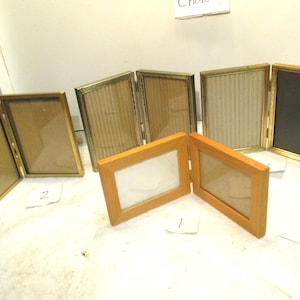 May include: A collection of hinged photo frames in various styles. Some have gold-colored metal frames, while others are made of wood. The frames are open, revealing the space for photos. The background is a white surface.