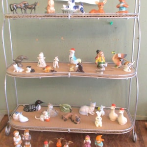 May include: A two-tiered metal shelf display filled with a variety of small ceramic figurines. The figurines depict animals, including a deer, a rooster, a cat, a dog, a duck, a loon, and a penguin. The shelf is labeled "Choice".