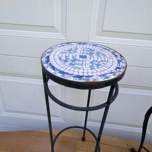 May include: A round, mosaic-tiled side table with a black wrought iron frame. The tabletop features a pattern of blue and white tiles. The table has a circular base and three legs, perfect for a patio or garden setting.