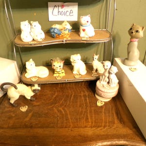 May include: A collection of ceramic cat figurines displayed on a tiered shelf and wooden table. The cats vary in size, color, and pose, including a Siamese cat, a cat with a heart, and a cat with a pink collar. The sign says "Choice".