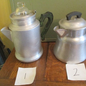 May include: Two vintage aluminum coffee pots. One pot has a clear glass top and a green handle. The other pot has a black handle. Both pots are sitting on a wooden surface. The number "1" is written on a white piece of paper next to the first pot, and the number "2" is written on a white piece of paper next to the second pot.