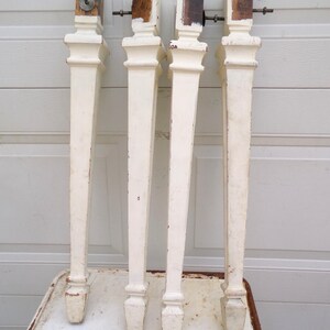 May include: Four tall, tapered furniture legs painted in an off-white finish with visible signs of wear and chipped paint. Each leg features a square top section with a metal bolt protruding from the side, a decorative molding, and a pointed foot.