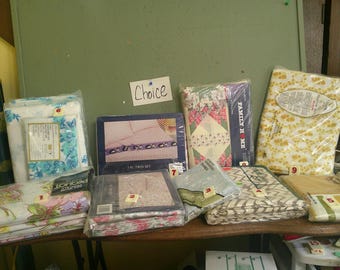 May include: A collection of nine packaged bedding sets, including sheets, pillowcases, and quilts, featuring various floral, geometric, and solid patterns. Each package is labeled with a numbered tag, and a small sign labeled Choice is visible.