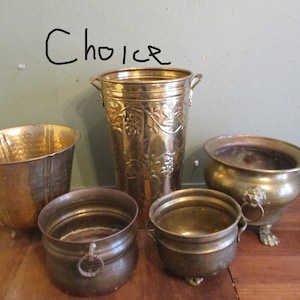 May include: A collection of antique brass planters and pots. The selection includes a hammered metal bucket, a tall vase with a grape vine design, and several small pots with handles and decorative feet. The word "Choice" is written on the wall.