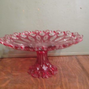 May include: A vintage, ruby red glass pedestal cake stand. The stand features a scalloped edge and a detailed, textured base. The glass is a vibrant red with clear accents, creating a striking contrast. This decorative piece is ideal for displaying desserts.