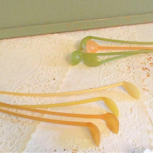 May include: A collection of vintage plastic cocktail stirrers in shades of orange, yellow, and green. The stirrers have a long, thin stem with a rounded or petal-shaped end. They are arranged on a white, lacy doily.