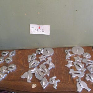 May include: A collection of vintage metal cookie cutters in various shapes, including hearts, leaves, animals, and geometric designs. The cutters are arranged on a wooden surface. The word "Choice" is written on a white note card.