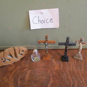 May include: A collection of religious items, including four crucifixes of varying sizes and materials, a small glass bottle, and a wooden fish with the word "Jesus" cut out. A note with the word "Choice" is pinned above.