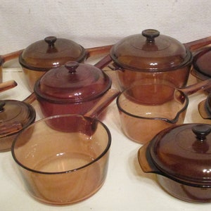 May include: A collection of amber-colored glass cookware, including saucepans with handles, pots with lids, and a casserole dish. The set is arranged on a white surface, showcasing the various sizes and shapes of the vintage kitchenware.