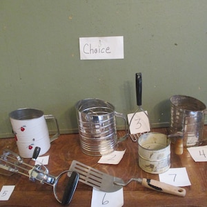 May include: A collection of vintage kitchen utensils, including a hand mixer, flour sifters, a potato masher, and a spatula. The items are arranged on a wooden surface, with a sign that says "Choice" in the background.