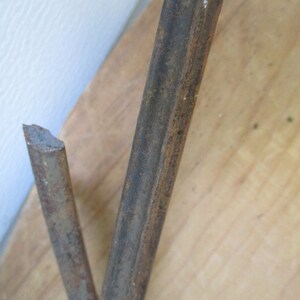 May include: Close-up of two rusty metal rods, one broken at an angle. The rods are brown and textured, with signs of corrosion. The background is a light-colored wooden surface and a white wall.