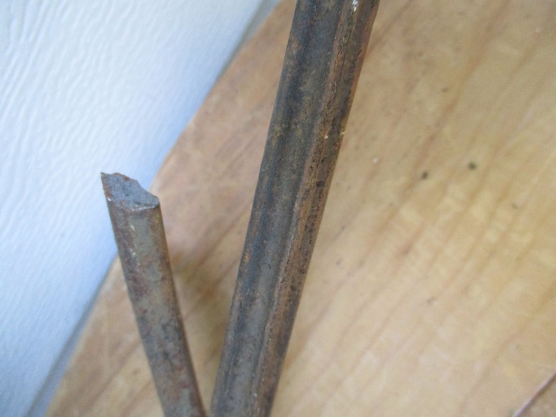 May include: Close-up of two rusty metal rods, one broken at an angle. The rods are brown and textured, with signs of corrosion. The background is a light-colored wooden surface and a white wall.