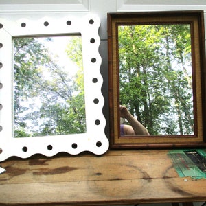 May include: Two decorative mirrors on a wooden table. One mirror has a white scalloped frame with circular cutouts. The other has a rectangular brown frame with a woven texture. Both reflect a green tree-filled scene.