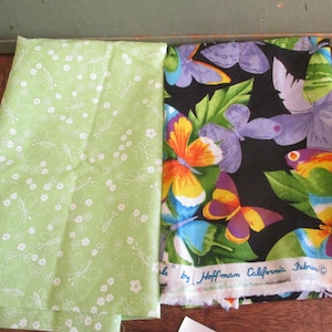 May include: Two pieces of fabric, one green with white butterfly designs and the other black with colorful butterfly designs. The black fabric has the text "Made by Hoffman California Fabrics" printed on it.