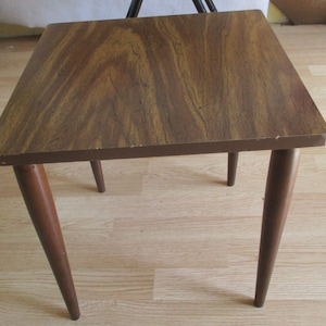 May include: A square wooden end table with a brown wood grain finish and four slender legs.