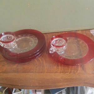 May include: Clear glass teacups and saucers with a red rim. The saucers have a circular design and a small indentation for the teacup. The teacups have a handle and a red rim. The set includes multiple saucers and teacups.