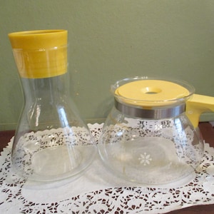 May include: Two vintage glass beverage containers with yellow lids. One is a carafe, the other a pitcher with a handle. Both have a clear glass body and a floral design. The carafe is approximately 9 inches tall.