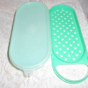 May include: An open, light green plastic container with a matching lid. The container has an oval shape with a perforated insert. The lid is smooth and solid. The container is designed for food storage.