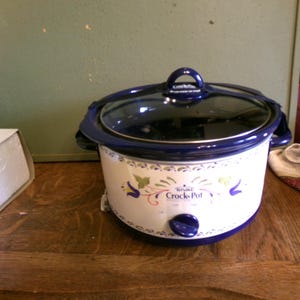 May include: A white and blue Rival Crock-Pot slow cooker with a glass lid. The slow cooker has a floral design and the words "Crock-Pot" are printed on the front. The slow cooker is sitting on a wooden surface.