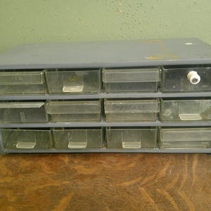 May include: A gray metal storage cabinet featuring twelve clear plastic drawers arranged in three rows of four. One drawer includes a white knob, and another drawer has the word "STANTART" written on the front. The unit has a rectangular frame.