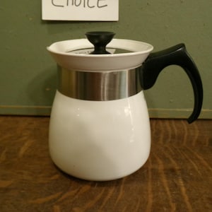 May include: A white teapot with a smooth, rounded body and a stainless steel band around the upper section. It features a black plastic handle and a matching black knob on the lid. The lid is white with a stainless steel rim, and the overall design is simple.