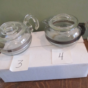 May include: Three glass teapots with brown and silver bands. The teapots are numbered 3 and 4 on white paper.