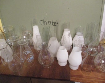 May include: A collection of various glass lamp shades and chimneys in clear, frosted, and white finishes. Some pieces feature blue floral patterns or etched designs. The word "chote" is written on the wall behind the assortment of glass items.