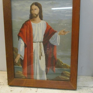 May include: Framed artwork depicting a figure in a white robe and red cloak, with long brown hair and a beard. The figure is set against a backdrop of water and rocks, framed in a wooden frame. The artwork is a painting.