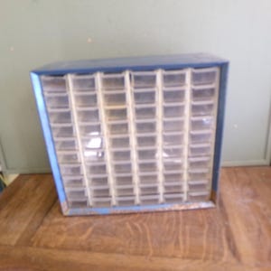 May include: A blue metal parts cabinet with numerous small, clear plastic drawers. The cabinet is rectangular and sits on a wooden surface. The drawers are arranged in a grid pattern, ideal for organizing small items.