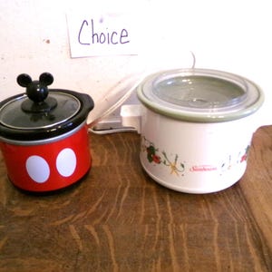 May include: Two small slow cookers on a wooden surface. One is red with white circles and a black lid with a Mickey Mouse handle. The other is white with a floral design and a clear lid.