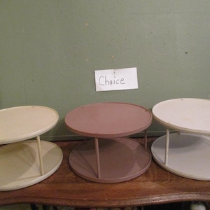 May include: Three oval, two-tiered display stands in cream, mauve, and off-white. Each stand has a matching base and top, connected by vertical supports. The stands are arranged on a wooden surface.