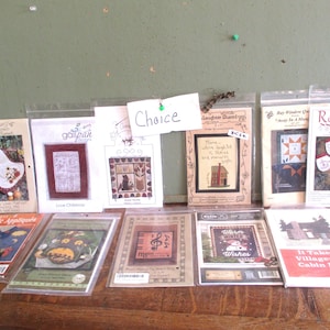 May include: A collection of 11 cross-stitch patterns featuring various themes, including winter, home, and nature. The patterns are displayed on a wooden surface with a sign that reads "Choice".