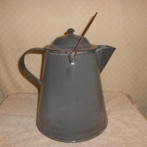 May include: A gray enamelware coffee pot with a lid and handle. The pot has a spout for pouring and a wire handle on the lid. The pot is sitting on a white surface.