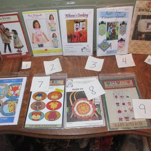 May include: A collection of sewing patterns and craft supplies, including patterns for aprons, baby bibs, and a sewing machine cover. The patterns feature images of children, animals, and various designs. The items are arranged on a wooden table.