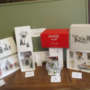 May include: Several Christmas village figurines and boxes are displayed on a wooden surface. The boxes feature illustrations of scenes with people and vehicles. A red box with the Coca-Cola logo is also present.