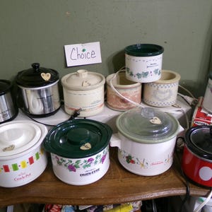 May include: A collection of ten small electric slow cookers in various colors and patterns, including white, red, and metallic finishes. Some feature floral or geometric designs, and one is red with white circles. A sign with the word Choice is visible.