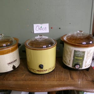 May include: Three slow cookers with glass lids are displayed on a wooden surface. The cookers are cream, yellow, and white with brown accents. The center cooker has the text "Choice" on a sign above it. The cookers have black knobs.