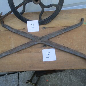 May include: An antique, rusty metal X-shaped piece, possibly a component of a larger mechanism, with curved ends. The number "3" is written on a white piece of paper. Other metal parts and a wheel are visible.