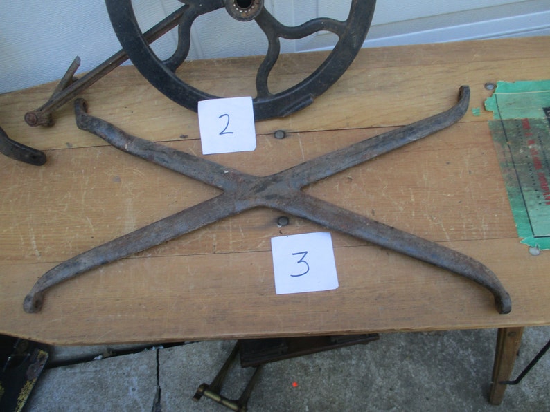 May include: An antique, rusty metal X-shaped piece, possibly a component of a larger mechanism, with curved ends. The number "3" is written on a white piece of paper. Other metal parts and a wheel are visible.