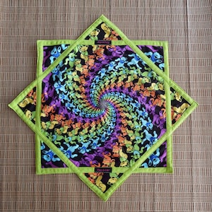 Puede incluir: Un salvamanteles cuadrado y acolchado con un fondo negro y un diseño en espiral de osos bailarines de colores. El salvamanteles está rematado con un borde verde brillante y una capa exterior verde en forma de estrella.