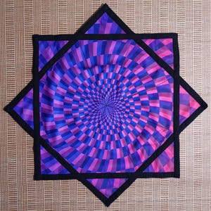 May include: A black-bordered, purple and blue patterned fabric square with a swirling, hypnotic design. The fabric is folded in a star shape.