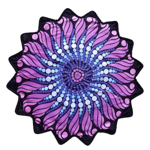 Puede incluir: Una alfombrilla circular con borde festoneado que presenta un fondo negro y un patrón central inspirado en un mandala. El diseño consiste en formas de pétalos púrpuras radiantes intercaladas con anillos concéntricos de puntos azul claro y blancos.