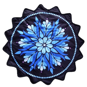 Puede incluir: Una alfombrilla de tela circular con borde festoneado, que presenta un fondo negro y un patrón floral central estilo mandala en tonos azules y blancos. El diseño incluye círculos concéntricos de puntos azul claro y pequeños acentos morados.