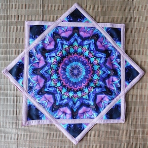 May include: A square, quilted coaster with a vibrant mandala design in shades of blue, purple, and pink. The coaster has a light pink border and is set against a neutral background. The design is symmetrical and detailed.