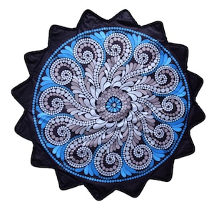 Puede incluir: Una alfombrilla circular con borde festoneado, fondo negro y un patrón de mandala central. El diseño consta de remolinos intrincados y pétalos radiantes en tonos azul brillante, azul claro, gris y blanco, creados con arte de puntos.