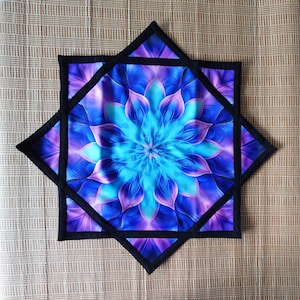 Puede incluir: Un edredón vibrante en forma de estrella con un diseño floral central en tonos azules y morados. El edredón tiene un borde negro y está sobre un fondo neutro. El diseño es simétrico y detallado.